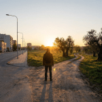 Person with backpack standing at junction of urban road and rural dirt path.
