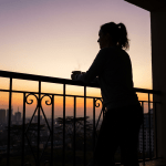 Woman silhouetted on a balcony at sunrise, holding a steaming mug.