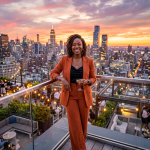 Woman in rust-colored suit holding cocktail on rooftop at sunset with city skyline