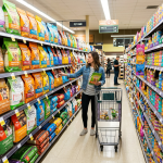 Woman in pet care aisle choosing dog food bag with shopping cart
