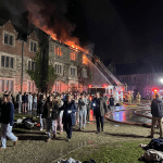Students walking outside a stone dormitory building with flames and smoke on the roof while firefighters spray water from a ladder truck.
